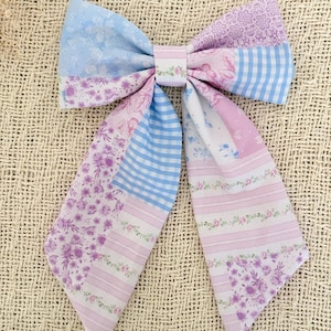Peut inclure: Un nœud pour cheveux en tissu présentant un motif patchwork dans des tons bleu clair, lavande et blanc. Le nœud comprend des sections à imprimés floraux, des carreaux vichy et des rayures horizontales, avec deux longs pans pointus.