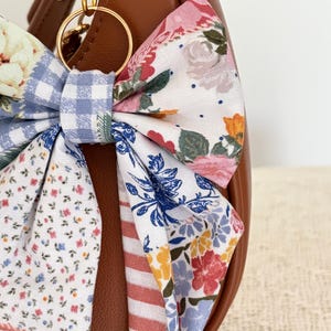 May include: A brown leather handbag featuring a large fabric bow attached to a gold-toned metal ring. The bow is constructed from various patterned fabrics, including blue and white gingham, small floral prints, and pink and white stripes.