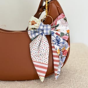 May include: A brown leather hobo bag featuring a decorative fabric bow keychain. The bow is constructed from various patterned fabrics, including blue gingham, floral prints, and pink stripes, attached to the bag with a gold-toned metal ring.