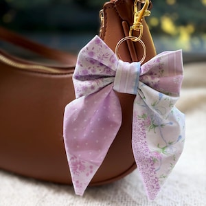 May include: A fabric bow keychain featuring a patchwork design in shades of lavender, light purple, and white. The bow displays various floral and polka dot patterns and is attached to a gold-toned metal lobster clasp and a circular ring.
