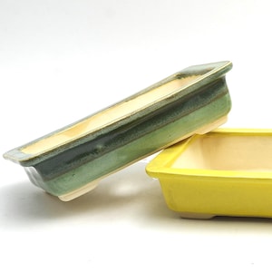 Può includere: Due vasi rettangolari in ceramica per bonsai. Uno è giallo vibrante, l'altro verde con un effetto sfumato. Entrambi hanno un interno di colore chiaro e sono rialzati su piccoli piedini. Ideale per piccole piante.