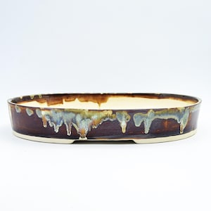Pode incluir: Um vaso oval de cerâmica para bonsai com perfil baixo e base plana. O exterior apresenta um esmalte castanho escuro com gotas decorativas em tons de bege claro e cinza-azulado que escorrem da borda. O interior é de cor creme claro.