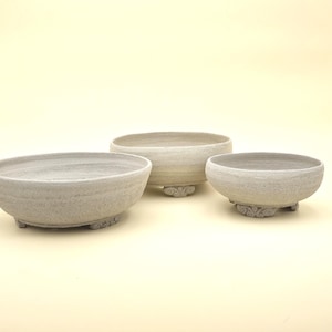 Peut inclure: Trois bols en céramique circulaires et peu profonds avec une finition gris clair texturée. Chaque bol présente un bord large et ouvert et repose sur trois petits pieds décoratifs avec un motif sculpté en spirale sous la base.