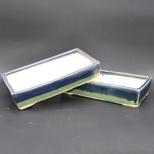Può includere: Due vasi rettangolari in ceramica per bonsai con interno bianco e smalto esterno bicolore, caratterizzati da una sezione superiore blu navy profondo e una sezione inferiore verde salvia chiaro. Ogni vaso ha un bordo piatto e piccoli piedini.