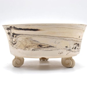 May include: A shallow, round ceramic bonsai pot featuring a cream-colored base with dark brown and black marbled, swirling patterns across the exterior. The vessel is supported by three small, curved, scroll-shaped feet at the base.