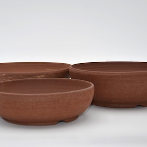 Peut inclure: Trois bols en céramique de couleur terre cuite de différentes tailles sont disposés sur une surface blanche. Les bols ont une forme arrondie avec une surface légèrement texturée. Les bols conviennent aux plantes ou à des fins décoratives.