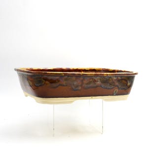 Può includere: Una fioriera rettangolare in ceramica con smalto marrone e crema. La fioriera ha un bordo leggermente rialzato ed è sostenuta da un supporto trasparente. Lo smalto ha un aspetto screziato con sfumature di marrone, verde e giallo.