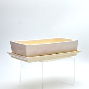 May include: A rectangular, pale pink ceramic planter with a matching rectangular base. The interior of the planter is a soft yellow. The planter and base are displayed on a clear acrylic stand. Suitable for small plants or succulents.