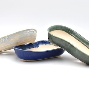 Peut inclure: Trois pots à bonsaï ovales en céramique avec de petits pieds. Un pot est gris clair avec un bord texturé, un est bleu roi profond avec une finition lisse, et un est vert forêt foncé avec un vernis brillant. Chaque pot a un intérieur crème.
