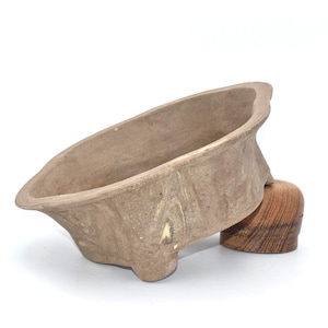 Op de afbeelding: Een ondiepe, ovaalvormige keramische kom met een getextureerde, matte beige afwerking en een brede, uitlopende rand. De kom rust op kleine, geïntegreerde voetjes en wordt aan één kant ondersteund door een afgerond, gepolijst houten blok.