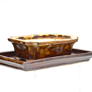 Könnte beinhalten: Ein rechteckiger Bonsai-Topf und die passende Schale. Der Topf ist in Braun- und Beigetönen glasiert und hat ein glänzendes Finish. Die Schale ist dunkler braun und ergänzt den Topf. Der Topf steht auf kleinen Füßen.