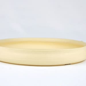 May include: A shallow, circular, cream-coloured ceramic dish with a smooth, matte finish and a slightly raised rim. Beside the dish stands a small, cylindrical brown cork stopper with printed text, providing a sense of scale for the ceramic piece.