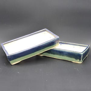 Może przedstawiać: Dwie prostokątne ceramiczne donice. Donice są glazurowane gradientem kolorów, w tym niebieskim, zielonym i białym. Donice mają niski profil i nadają się do bonsai lub małych roślin.