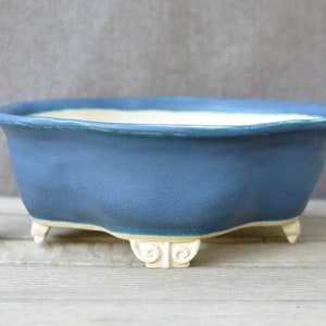 May include: A blue ceramic bonsai pot with a matte finish. The pot has a wide, oval shape and sits on three small, decorative feet. A cork is next to the pot.
