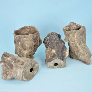 Puede incluir: Cuatro maceteros irregulares con aspecto de roca y acabado jaspeado en tonos marrón y gris. Cada recipiente cuenta con un centro hueco y un orificio de drenaje en la base, mostrando una textura rugosa y natural con formas orgánicas.