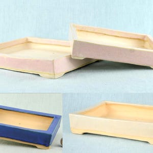 Może przedstawiać: Trzy prostokątne ceramiczne doniczki do bonsai z niskimi ściankami i małymi nóżkami. Dwie doniczki są jasnoróżowe z kremowym wnętrzem, a jedna jest ciemnoniebieska z kremowym wnętrzem. Obok znajduje się mały cylindryczny korek.