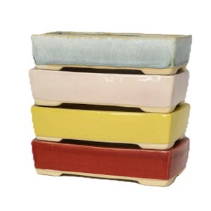 May include: A stack of four rectangular bonsai pots in pastel colors: light blue, pale pink, yellow, and red. Each pot has a slightly textured surface and sits on small, rounded feet. The pots are stacked neatly, showcasing their varied hues.