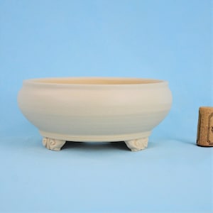 May include: A white ceramic bonsai pot with a rounded oval shape and three decorative feet. The pot is sitting on a light blue surface.