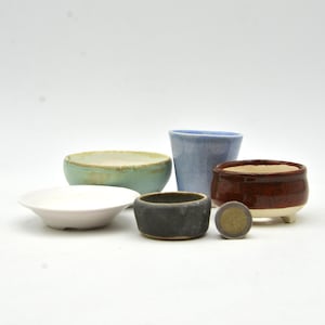 Peut inclure: Une collection de petits pots en céramique de différentes formes et couleurs. Comprend un bol peu profond blanc, une tasse bleu clair, un pot gris foncé, un bol turquoise et un pot marron avec des pieds blancs. Sur fond blanc.