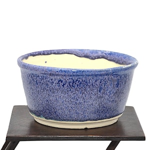May include: A round ceramic bowl featuring a deep blue and violet mottled glaze on the exterior, with a smooth cream-coloured interior and a thick, uneven rim. The base of the bowl is unglazed, revealing a light beige ceramic material.
