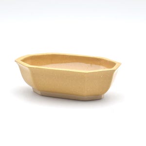 Puede incluir: Un cuenco de cerámica octogonal y alargado con un acabado esmaltado en color amarillo claro. El recipiente presenta una parte superior abierta y ancha con lados inclinados que se estrechan hacia una base plana más pequeña, de color sólido.