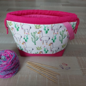 Könnte beinhalten: Eine Stofftasche mit weißem Hintergrund und einem wiederkehrenden Muster aus Lamas und grünen Kakteen, akzentuiert durch einen leuchtend pinken oberen Rand, Kordelzug und Tragegriff. Ein Knäuel mehrfarbiges Garn und fünf Stricknadeln aus Holz liegen bei.
