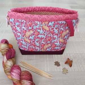 Könnte beinhalten: Eine Stofftasche für Handarbeitsprojekte mit einem Muster aus Eichhörnchen und Eicheln in Rosa und Hellblau, mit dunkelweinrotem Boden, rosa Kordelzug und braun gepunktetem Innenfutter, dazu ein Strang mehrfarbiges Garn und Holzstricknadeln.