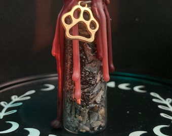 May include: A small glass bottle filled with dried herbs and topped with a red wax seal. A gold paw print charm hangs from the bottle.