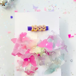 May include: A white card features a purple barrette with the gold numbers "1989". Below, a large hair bow is adorned with pink, purple, and blue butterflies. The bow is made of sheer fabric and has a glittery appearance. The background is white with confetti.