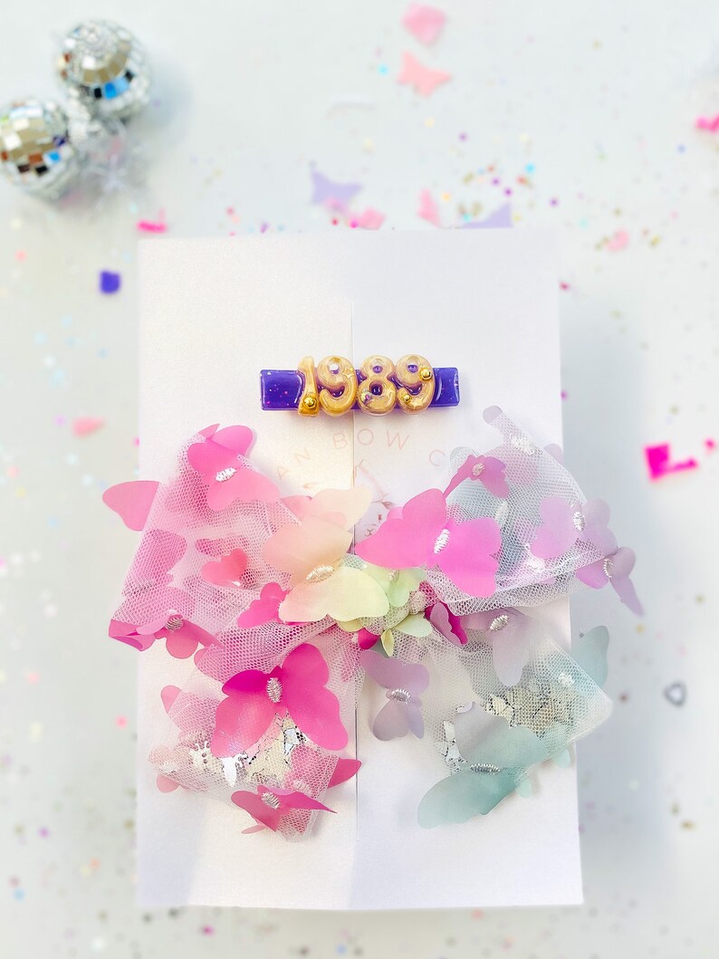May include: A white card features a purple barrette with the gold numbers "1989". Below, a large hair bow is adorned with pink, purple, and blue butterflies. The bow is made of sheer fabric and has a glittery appearance. The background is white with confetti.