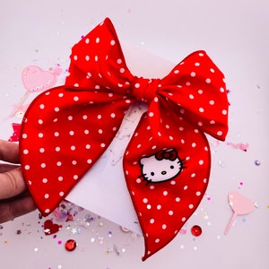 May include: A large red bow with white polka dots and a Hello Kitty graphic. The bow is made of a shiny fabric and is tied in the center. The Hello Kitty graphic is on one of the bow's tails. The bow is on a white background.