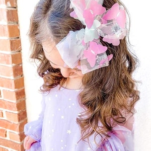 May include: A large hair bow featuring pink and white butterfly accents. The bow is attached to the hair of a person wearing a lavender dress with a star pattern. The bow is made of a sheer material.