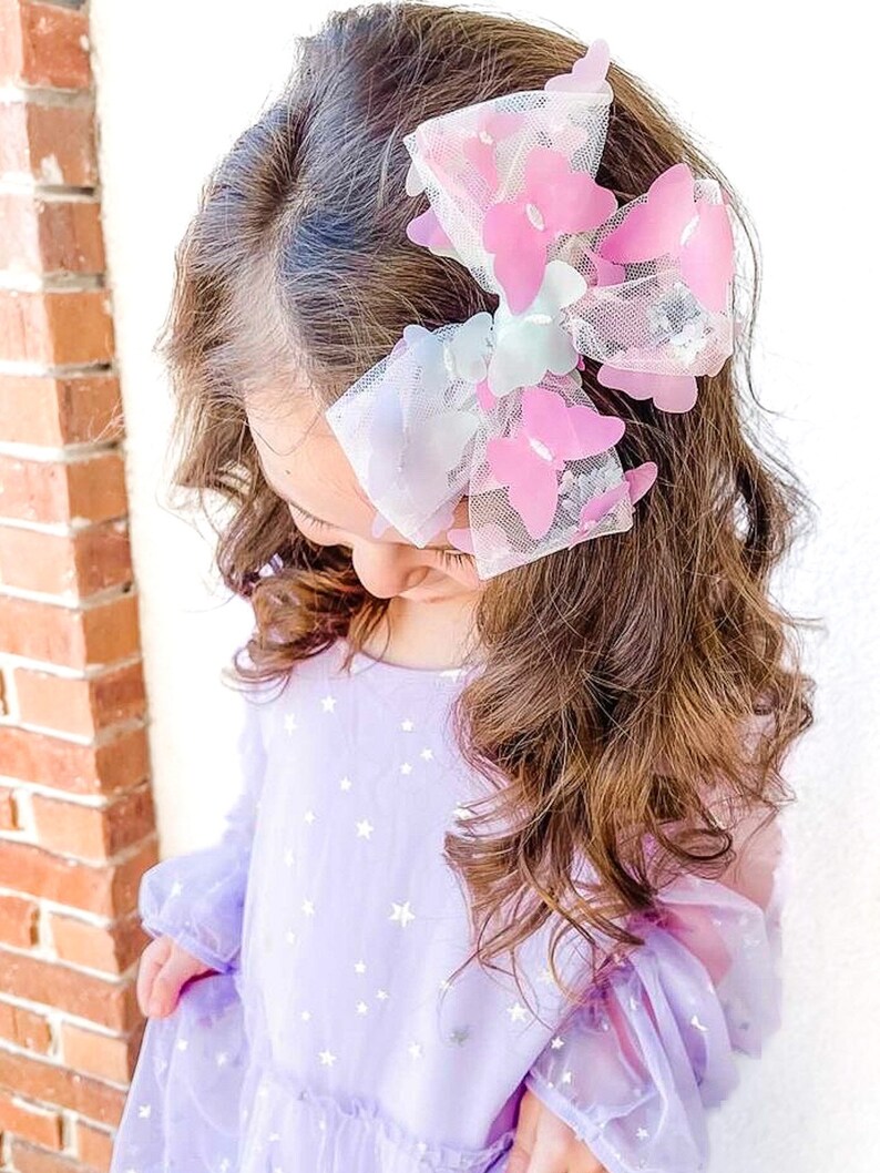 May include: A large hair bow featuring pink and white butterfly accents. The bow is attached to the hair of a person wearing a lavender dress with a star pattern. The bow is made of a sheer material.
