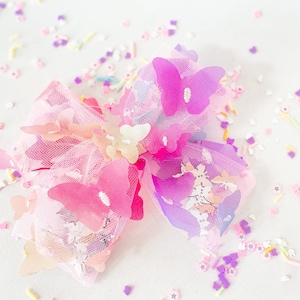 May include: A large, decorative hair bow featuring layers of pink tulle and adorned with multiple butterfly accents in shades of pink and purple. The bow is embellished with small, colorful sprinkles and flower-shaped confetti.