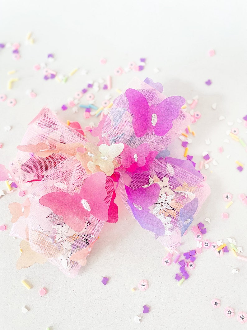 May include: A large, decorative hair bow featuring layers of pink tulle and adorned with multiple butterfly accents in shades of pink and purple. The bow is embellished with small, colorful sprinkles and flower-shaped confetti.