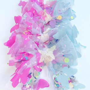 May include: Two decorative hair bows. One bow is pink and the other is blue. Both bows are made of tulle and decorated with sequins and butterfly-shaped embellishments. The bows are on a white background.