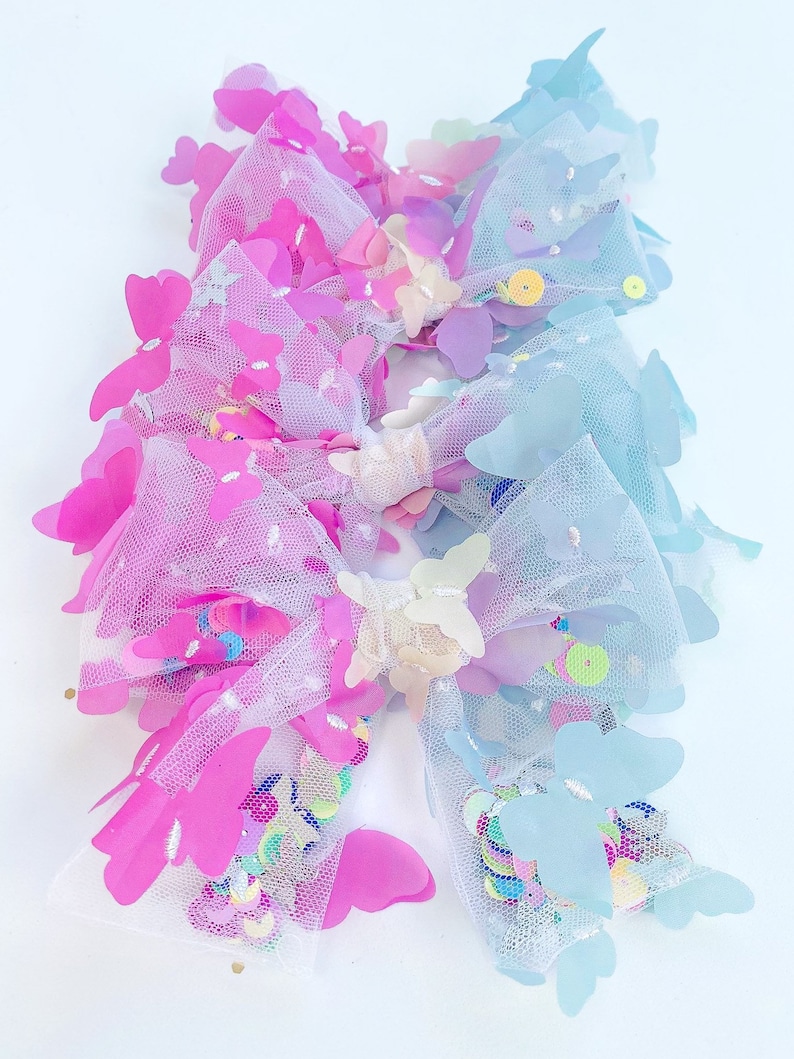 May include: Two decorative hair bows. One bow is pink and the other is blue. Both bows are made of tulle and decorated with sequins and butterfly-shaped embellishments. The bows are on a white background.