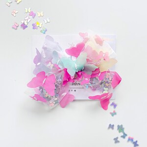 May include: A colorful hair bow featuring pink, blue, and yellow butterfly accents. The bow is made of tulle and adorned with small, iridescent butterfly sequins. The bow is on a white card with the word "eanbow" visible.