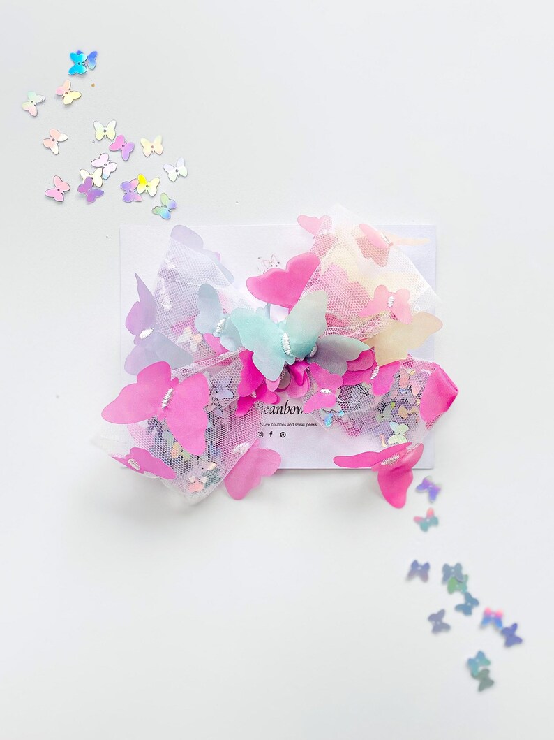 May include: A colorful hair bow featuring pink, blue, and yellow butterfly accents. The bow is made of tulle and adorned with small, iridescent butterfly sequins. The bow is on a white card with the word "eanbow" visible.