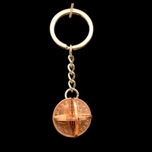 Könnte beinhalten: Ein kupferfarbener Schlüsselanhänger mit einem goldfarbenen Ring und einer Kette. Der Anhänger ist eine Münze mit einem kreuzförmigen Design. Die Münze wird von einem kupferfarbenen Metallband zusammengehalten. Der Hintergrund ist schwarz.