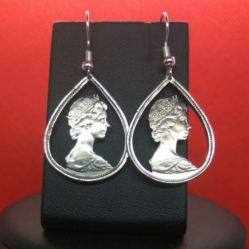 May include: A pair of silver teardrop-shaped earrings with a profile of a woman's face cut out of the center. The earrings are hanging from a black display stand.