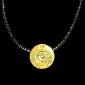 Puede incluir: Un collar con colgante dorado con un diseño de casquillo de bala. El colgante circular tiene texto grabado y una hendidura central. El collar tiene un cordón negro. El fondo es negro.
