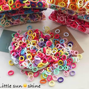 May include: A collection of colorful hair ties in various shades, including pink, yellow, blue, and green. Some are scattered on a plate, while others are in clear plastic bags. The text "Little sun shine" is at the bottom.