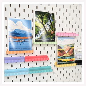 May include: A white pegboard wall features multiple colorful, wavy-edged plastic shelves holding three national park prints. The prints depict Kobuk Valley, Shenandoah, and Theodore Roosevelt National Parks. The shelves are orange, light green, pink, purple, red, light blue, teal, yellow, white, and black.
