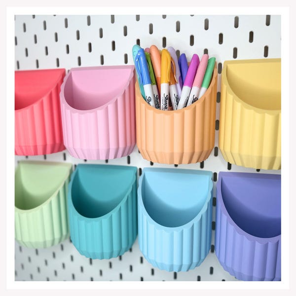 May include: A set of eight colorful, ribbed, semi-circular wall-mounted storage cups attached to a white pegboard. The cups are in shades of red, pink, orange, yellow, light green, teal, light blue, and purple. One orange cup holds several Sharpie markers.