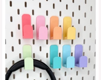 May include: A set of seven U-shaped wall hooks in various pastel colours including coral, pink, orange, yellow, light green, turquoise, blue, and purple, attached to a white pegboard, with a black headband resting on the light green hook.