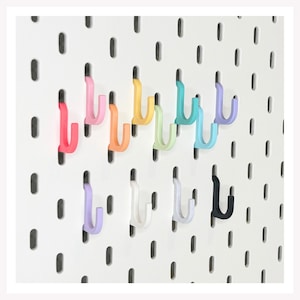 May include: A collection of J-shaped hooks in various colors including pink, orange, yellow, light green, teal, light blue, purple, white, grey, and black, attached to a white pegboard with vertical oval slots for organization and storage.