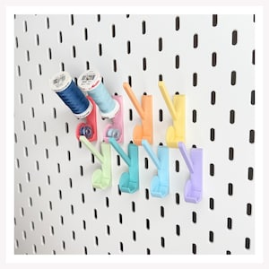 May include: A white pegboard with a grid of vertical slots features several colorful plastic hooks attached to it. Two spools of sewing thread in dark blue and light blue are placed on the top two hooks, while the remaining hooks are empty.