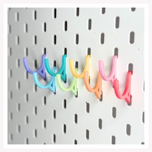 May include: A series of U-shaped hooks in a rainbow gradient of colors, including purple, blue, green, yellow, orange, and pink, attached to a white pegboard with vertical slots. The hooks are arranged in a horizontal row across the board.
