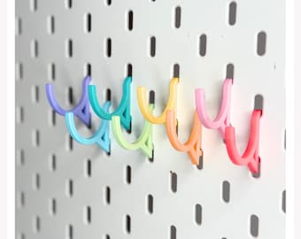 Könnte beinhalten: Eine Reihe von U-förmigen Haken in einem Regenbogen-Farbverlauf aus Violett, Blau, Grün, Gelb, Orange und Pink, die an einer weißen Lochplatte mit vertikalen Schlitzen befestigt sind. Die Haken sind in einer horizontalen Reihe angeordnet.